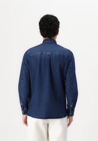 Camisa de mezclilla azul oscuro con mangas largas, puños abotonados y una yugo en la parte posterior. Presenta un cuello puntiagudo y un dobladillo recto en la parte inferior.