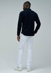 Hombre de pie de espaldas, vistiendo una camiseta negra de manga larga, jeans blancos y zapatillas blancas, frente a un fondo liso.