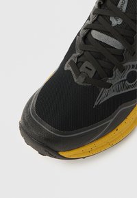 Ténis desportivo preto com parte superior em malha texturizada, detalhes cinzentos e sola amarela com manchas. Possui atacadores pretos e biqueira reforçada.