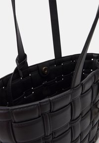 Sac fourre-tout en cuir tissé noir avec deux longues anses. Surface texturée présentant un motif de panneaux entrelacés et une fermeture magnétique.
