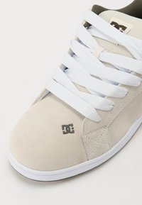 Világos bézs velúr sneaker fehér fűzőkkel, kerek orral és szürke logóval. Továbbá, varrott díszítésekkel és fehér gumitalppal rendelkezik.