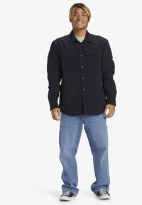 Schwarzes Hemd mit Knöpfen und zwei vorderen Taschen, getragen über einem hellgrünen Hemd, kombiniert mit hellblauen Jeans und schwarzen Sneakers.