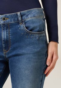 Jeans de mezclilla azul con un corte clásico, que presentan remaches plateados decorativos en el bolsillo delantero. La tela tiene un ligero efecto de lavado y una textura suave.