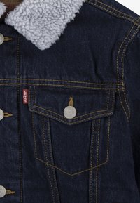 Chaqueta de mezclilla azul oscuro con un cuello de forro polar gris, que presenta botones de metal plateado, dos bolsillos en el pecho, costuras amarillas y el logo de la marca.