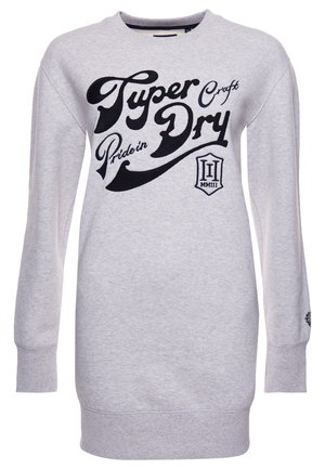Vrouw draagt een groene doorgestikte jas over een grijze sweatshirtjurk met zwarte letters, beige sokken en witte sneakers, staand binnen.