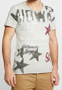 Camiseta de algodón gris con cuello en V, con patrones de estrellas negras y rojas, impresiones de texto y detalles desgastados. Manga corta y ajuste relajado.