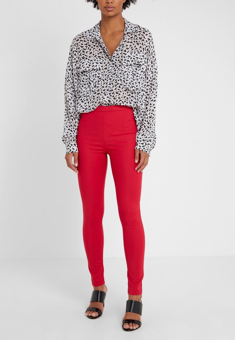 Blouse blanche à motifs abstraits noirs, manches longues, et deux poches poitrine. Leggings rouges taille haute et sandales noires à talons carrés.