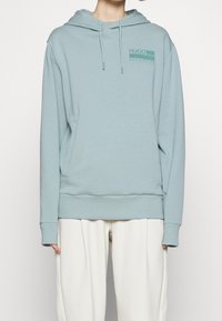 Sudadera azul claro con bolsillo frontal, capucha ajustable con cordón, puños acanalados y un logo gráfico en azul más oscuro. Material de algodón suave.