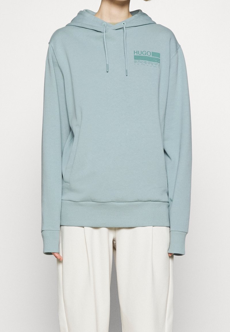 Sudadera azul claro con bolsillo frontal, capucha ajustable con cordón, puños acanalados y un logo gráfico en azul más oscuro. Material de algodón suave.