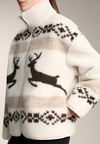 Biała kurtka z polaru z brązowymi wzorami reniferów i wzorami geometrycznymi, z wysokim kołnierzem i pełnym zamkiem błyskawicznym. Miękka, teksturowana powierzchnia z kontrastowymi kolorami.