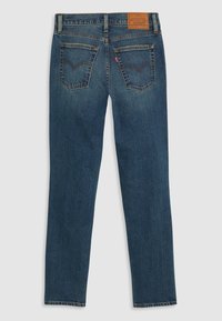 Jean en denim bleu foncé avec poches arrière, écusson logo en cuir marron et étiquette rouge sur la poche droite, vu de dos.