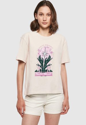 Lyserød t-shirt med korte ærmer, der har et grafik af mørke hænder, som holder blomster og en stjerneblomst, med teksten "SKINNELYSE" ovenfor.