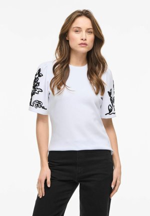 Kobieta o długich brązowych włosach, ubrana w biały krótki rękaw z czarnym kwiatowym haftem na rękawach i czarne spodnie, stojąca na białym tle.