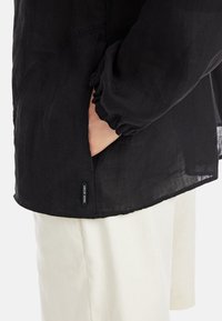Schwarze, leichte Bluse mit langen Ärmeln und gerafften Manschetten, mit einem seitlichen Schlitz und einem kleinen Etikett am Saum. Getragen über weißen Hosen.