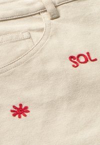 Beige stof met twee rode geborduurde details: het woord "SOL" en een stralenpatroon. De textuur is glad, afgebeeld vanuit een hoek.