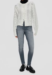 Cárdigan gris claro de punto con un patrón texturizado, cuello redondo y cierre con botones, combinado con vaqueros ajustados gris oscuro y zapatos negros.