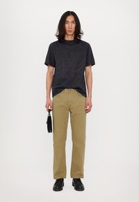 Tricou negru cu mânecă scurtă și model subtil, asortat cu pantaloni bej drepti. Modelul ține o geantă neagră și poartă pantofi negri.
