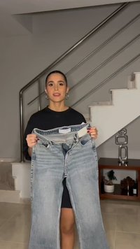 Γυναίκα με μαλλιά δεμένα προς τα πίσω, που στέκεται σε εσωτερικό χώρο, κρατώντας ένα ζευγάρι ανοιχτό μπλε τζιν μπροστά από μια μεταλλική σκάλα και ένα ράφι με διακοσμητικά.