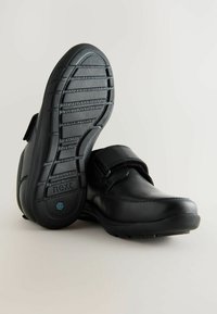 Chaussures en cuir noir avec une bride Velcro, bout rond et semelle en caoutchouc texturé avec des rainures horizontales pour une meilleure adhérence. Étiquette de marque sur la semelle.