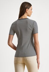 Sieviete, kas valkā īsroku zilu ar baltām svītrām krekliņu un bēšas krāsas bikses, stāvot ar muguru pret kameru uz vienkrāsaina baltā fona.