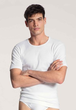 Weißes Kurzarm-T-Shirt mit runder Halslinie, aus weichem Stoff, figurbetont, getragen mit verschränkten Armen vor einem hellen Hintergrund.