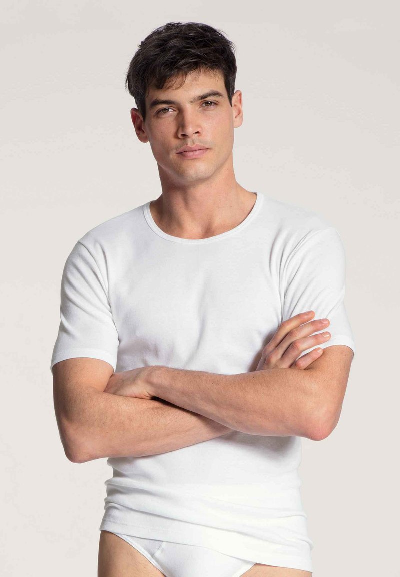 Weißes Kurzarm-T-Shirt mit runder Halslinie, aus weichem Stoff, figurbetont, getragen mit verschränkten Armen vor einem hellen Hintergrund.