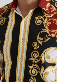 Chemise à col avec des motifs baroques noirs, blancs, rouges et dorés, ornée de rayures verticales et de tourbillons décoratifs, fermée par des boutons blancs.