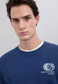 Sudadera azul marino con cuello redondo, que presenta un logo gráfico en azul claro de un balón de baloncesto y el texto "Indoors & Outdoors Scalpers Co."