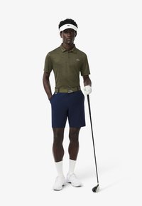 Polo verde oliva con un patrón sutil, pantalones cortos azules marino, zapatos blancos y visera; sosteniendo un palo de golf; atuendo atlético casual, diseño funcional.