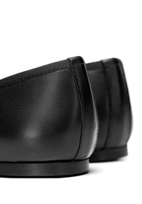 Zapatos de cuero negro con una textura suave, parte trasera redondeada y perfil bajo. Presentan un detalle de costura sutil y una suela plana.