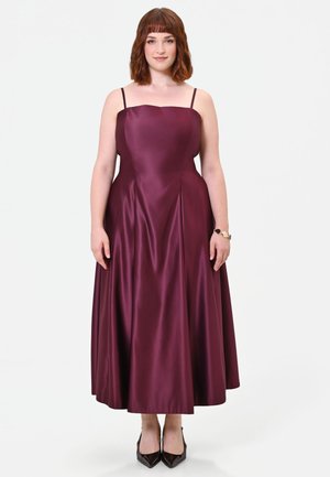 Donna con capelli fino alle spalle che indossa un abito midi in raso burgundy senza maniche e tacchi neri a punta, in piedi su uno sfondo bianco.