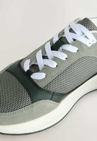 Šedá sportovní bota vyrobená z prodyšné síťoviny a semiše, s bílými tkaničkami, tónovými zelenými akcenty a bílou podrážkou s zdrsněným vzorkem.