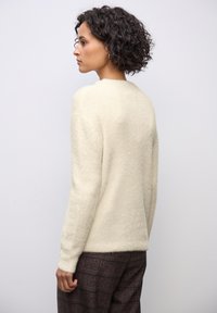 Maglione color crema con una finitura morbida e texture, caratterizzato da maniche lunghe e una vestibilità rilassata, abbinato a pantaloni a quadri marroni.