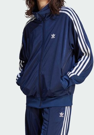 Persona vistiendo un chándal azul marino de Adidas con franjas blancas y logo, con las manos en los bolsillos de la chaqueta, de pie frente a un fondo claro y liso.