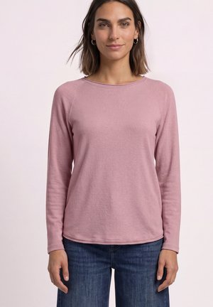 Femme aux cheveux bruns mi-longs, portant des boucles d’oreilles anneaux, un haut rose à manches longues et un jean bleu, debout devant un fond clair uni.