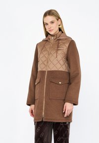 Cappotto marrone trapuntato con cappuccio, dotato di un pannello trapuntato diagonale, due tasche frontali e una cerniera completa. Texture del tessuto morbida.