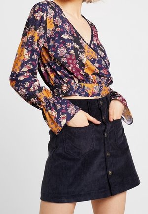 Kvinde iført en blomster-mønstret langærmet slå-om-bluse i navy, sennep og bordeaux med en sort kordnørlangs foran nederdel, hænder i lommer.