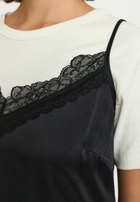 Mustavilla satiininen hihaton toppi pitsireunalla valkoisen T-paidan päällä; toppi on sileäpintainen ja siinä on säädettävät olkaimet.