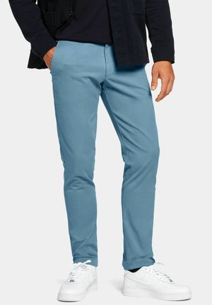 Man die lichtblauwe, slim-fit broek, een zwart jack en witte sneakers draagt, staand met één hand in de zak.