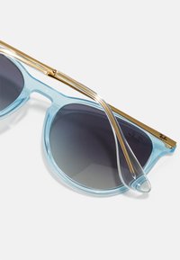 Ochelari de soare bleu deschis cu lentile gri cu gradient, un cadru transparent și un accent din metal aurit pe sprânceană. Prezintă o formă elegantă și rotunjită.