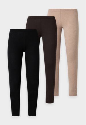Femme aux cheveux tressés, portant un t-shirt blanc ajusté à manches courtes, des leggings noirs et des baskets blanches, sur un fond blanc uni.