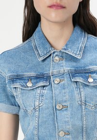 Veste en denim bleu clair à manches courtes, avec des boutons-pression et deux poches avant. Présente une texture délavée et des détails de couture contrastés.