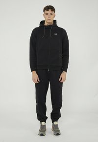 Haut à fermeture éclair noir avec poches avant et capuche réglable, associé à un pantalon de survêtement noir et des baskets multicolores. Texture lisse tout au long.