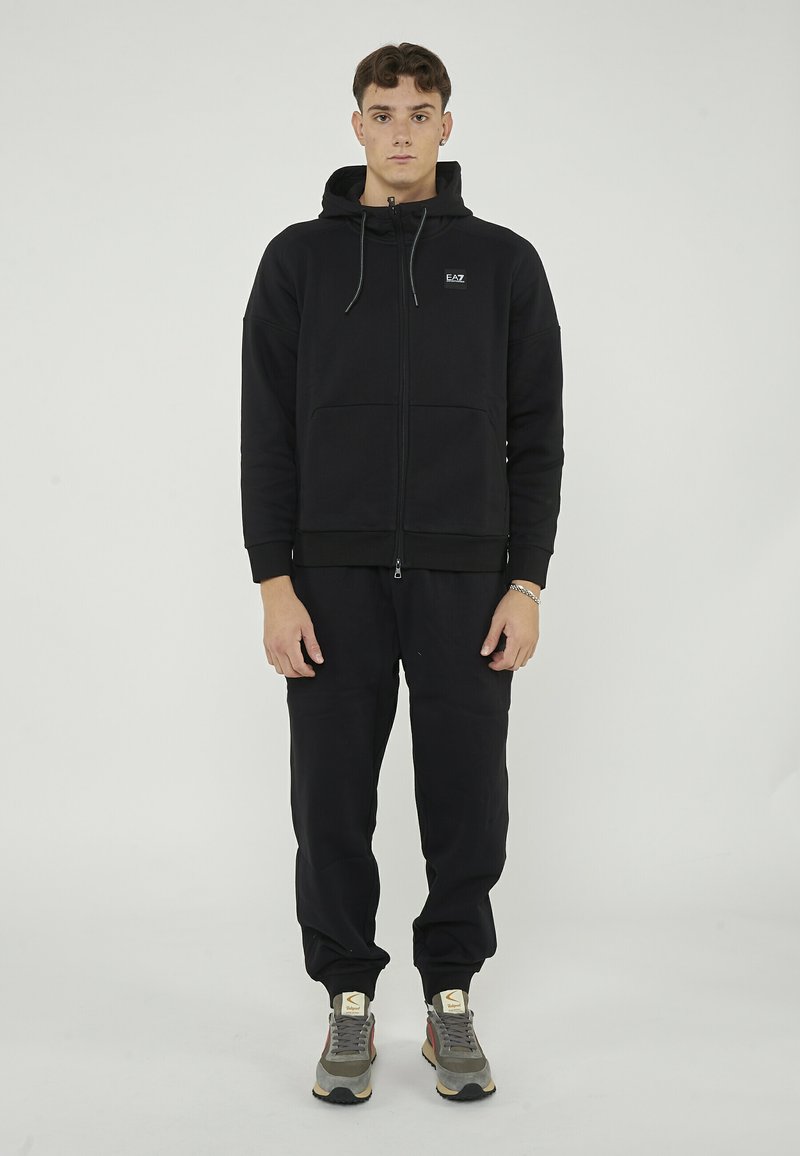 Haut à fermeture éclair noir avec poches avant et capuche réglable, associé à un pantalon de survêtement noir et des baskets multicolores. Texture lisse tout au long.