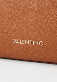 Hnědá kabelka z texturované kůže s zlatým logem "Valentino" na přední straně, zdůrazňující její sofistikovaný design a kvalitu materiálu.