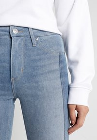 Close-up of a person wearing light blue denim jeans and a white long-sleeve top, with their right hand relaxed by their side.