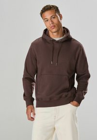 Brauner Baumwoll-Hoodie mit verstellbaren Kordeln, Fronttasche, gerippten Bündchen und Saum; getragen mit hellen Hosen.