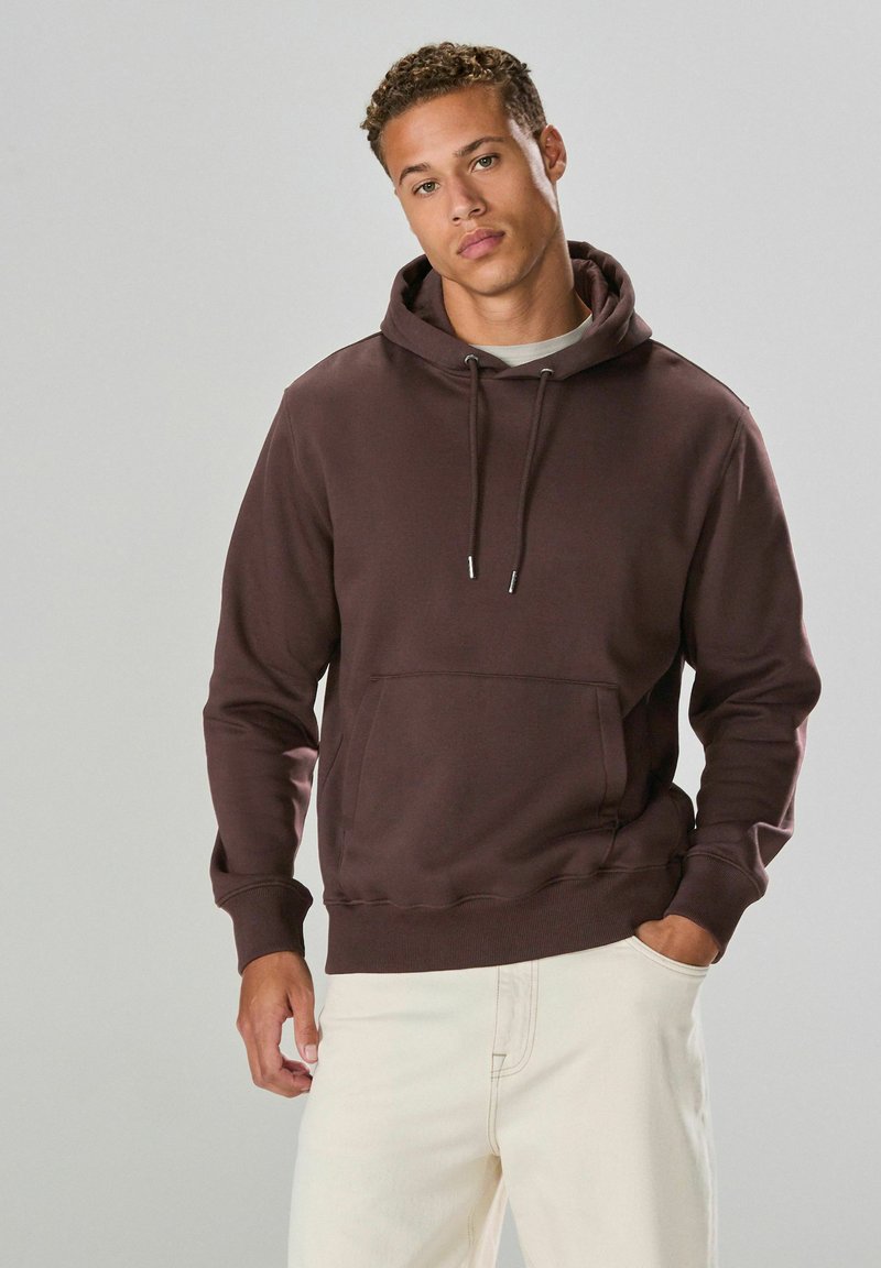 Brauner Baumwoll-Hoodie mit verstellbaren Kordeln, Fronttasche, gerippten Bündchen und Saum; getragen mit hellen Hosen.