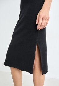 Μαύρο πλεκτό φόρεμα με πλαϊνό σκίσιμο, ριγωτή υφή και εφαρμοστή σιλουέτα. Κοντινό πλάνο χεριού που ξεκουράζεται κοντά στο σκίσιμο.