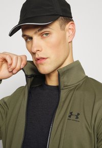 Olivgrüne Zip-Jacke mit hohem Kragen, schwarzem Logo und schwarzem T-Shirt darunter. Schwarze Mütze mit weißem Rand. Die Hand des Models hält den Kragen.
